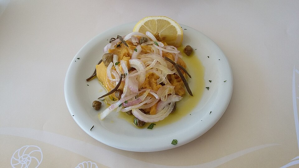A bowl of smooth yellow split pea puree topped with raw red onion, capers and olive oil