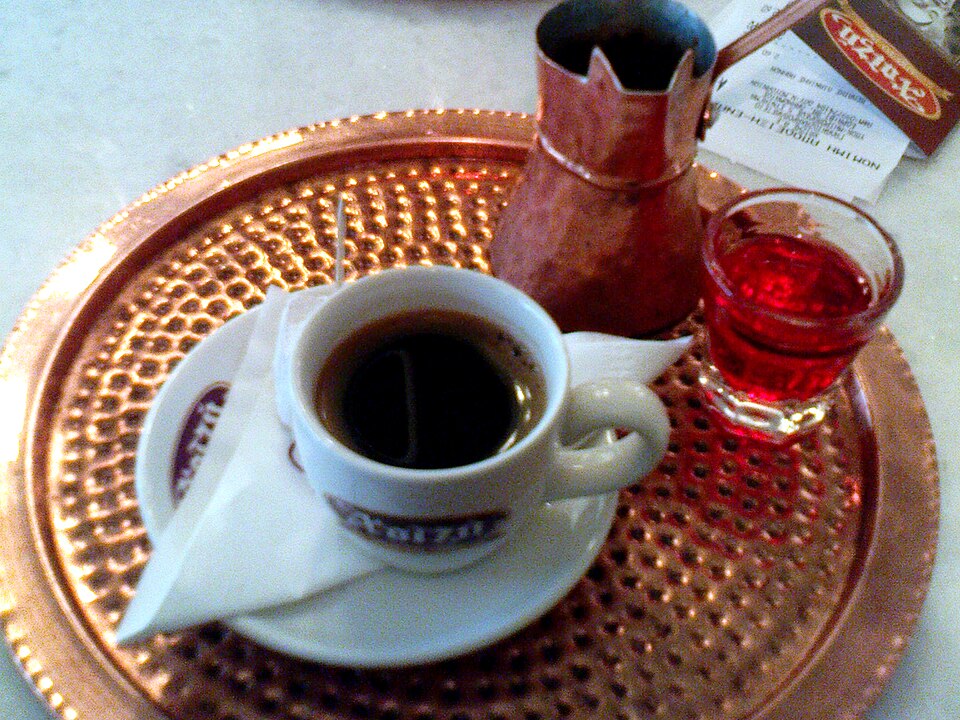 A small cup of Greek coffee with thick foam on top beside a copper briki