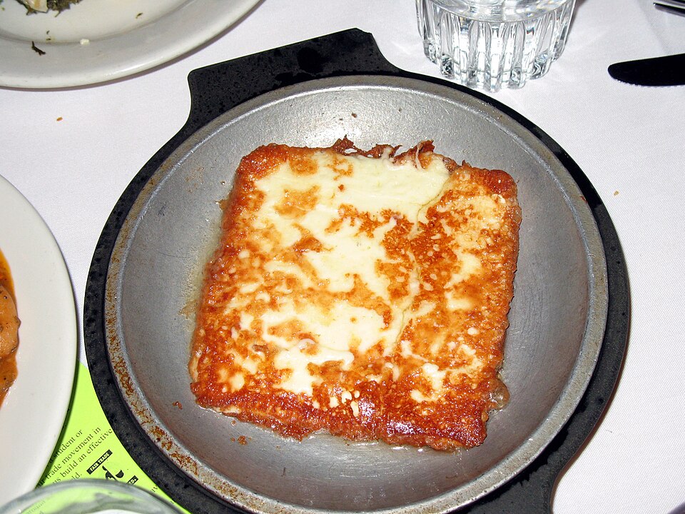 A slice of golden fried Greek cheese in a small pan with a lemon wedge